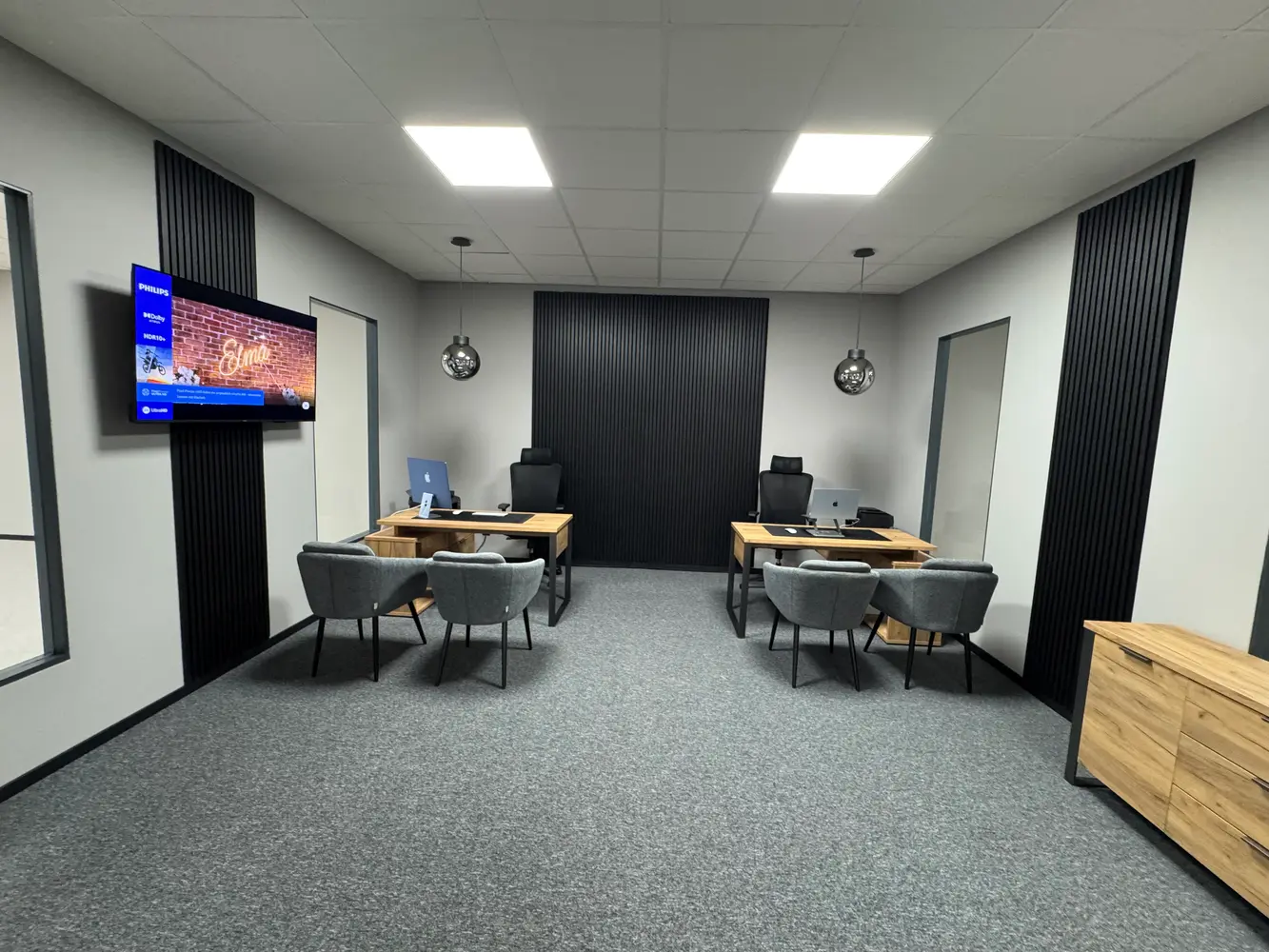 Ein modernes Büro mit zwei Schreibtischen, grauen Stühlen und einem Fernseher an der Wand