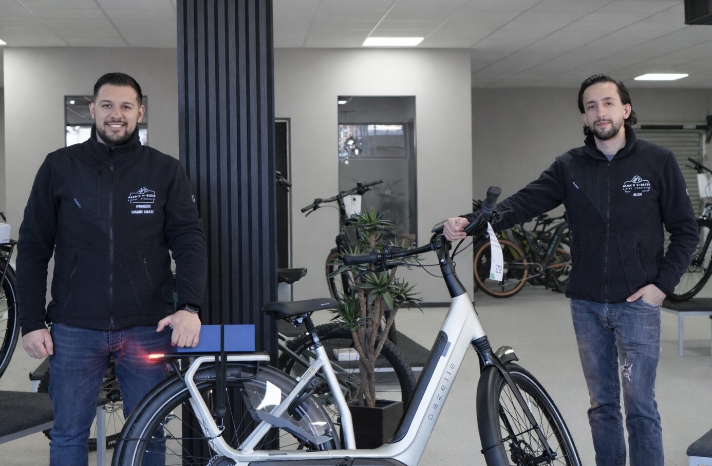 Zwei Männer in dunkler Kleidung stehen freundlich lächelnd neben einem weißen Fahrrad